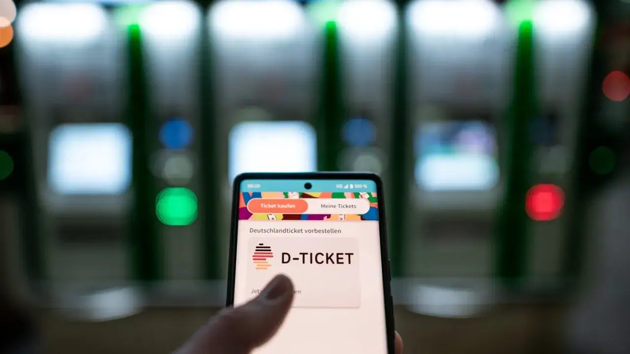 Smartphone zeigt "D-TICKET" an, bereit für das Deutschlandticket per Lastschrift. Im Hintergrund unscharfe Automaten.