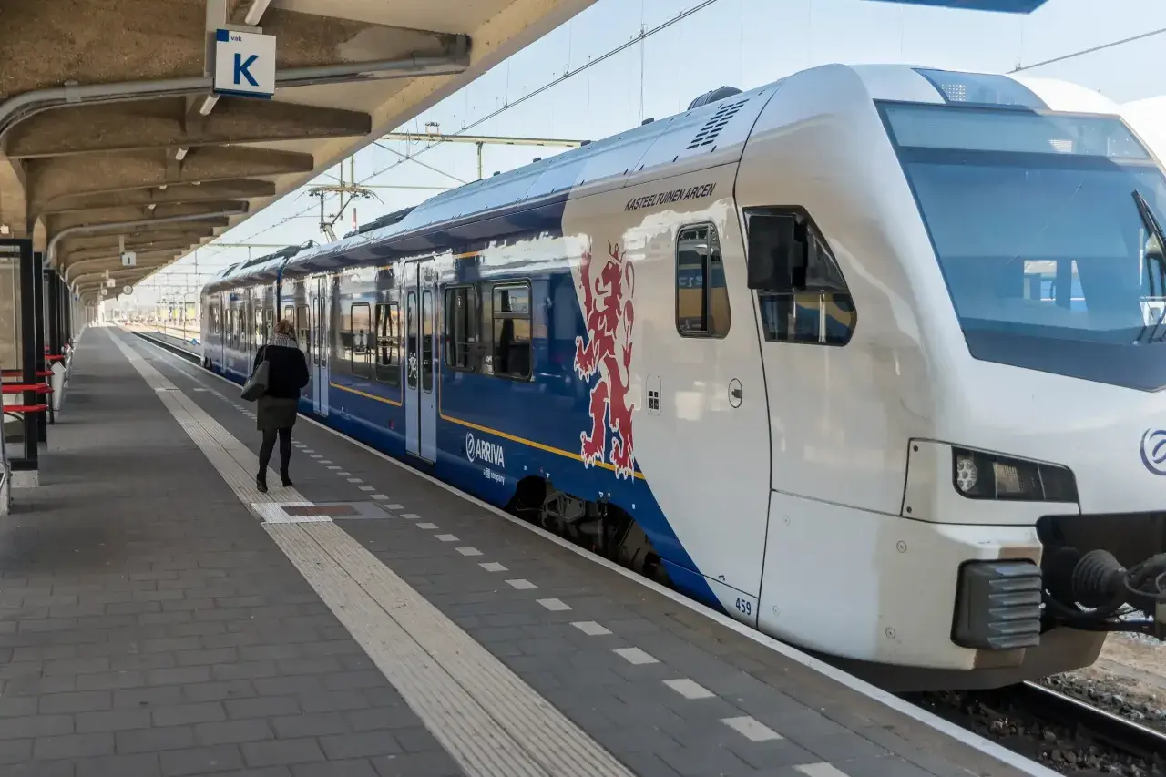 Eine Frau wartet auf dem Bahnsteig auf den Zug. Der blaue Arriva-Zug mit dem roten Löwenwappen ist ein Teil des Nahverkehrs in den Niederlanden.