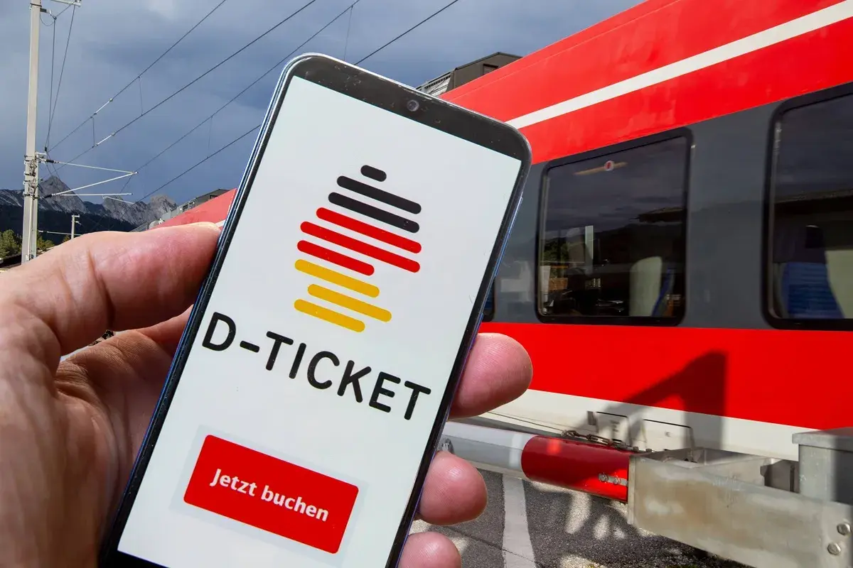 Smartphone zeigt D-Ticket App mit "Jetzt buchen" Button. Im Hintergrund rote Bahn in NRW.
