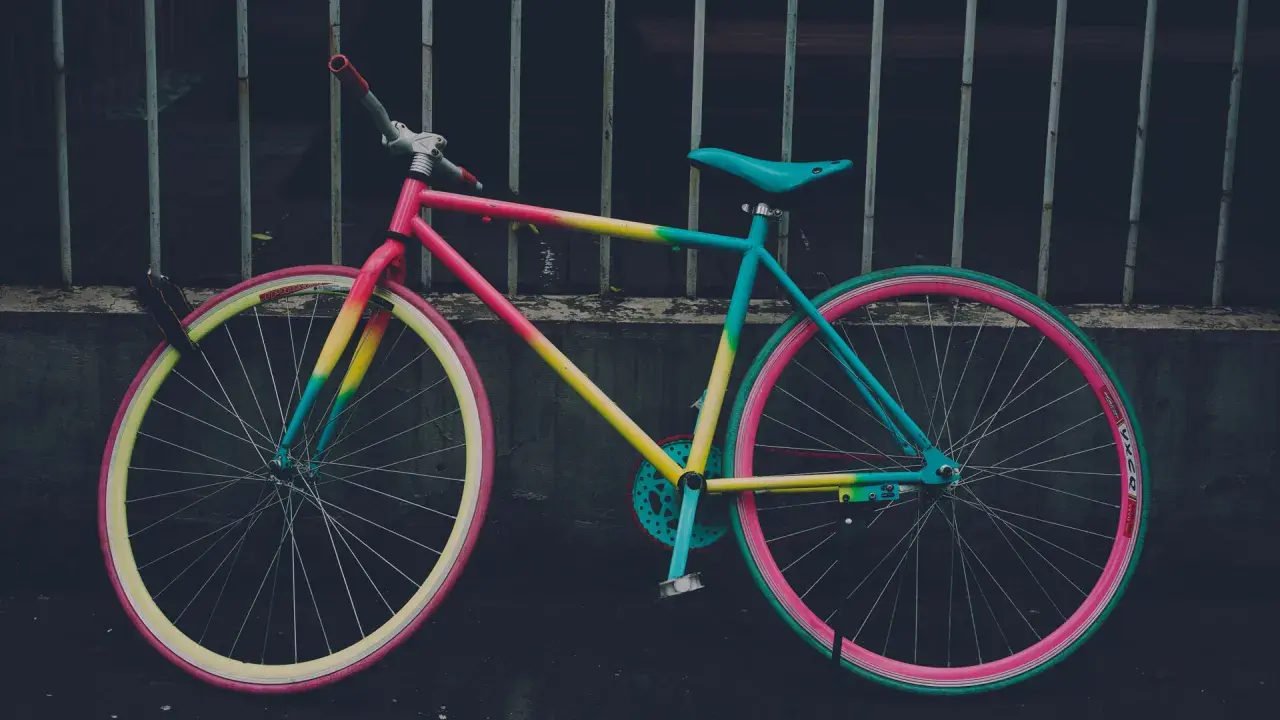 Ein bunt lackiertes Fahrrad mit pinken, gelben und türkisen Akzenten, das an einem Gitter lehnt.