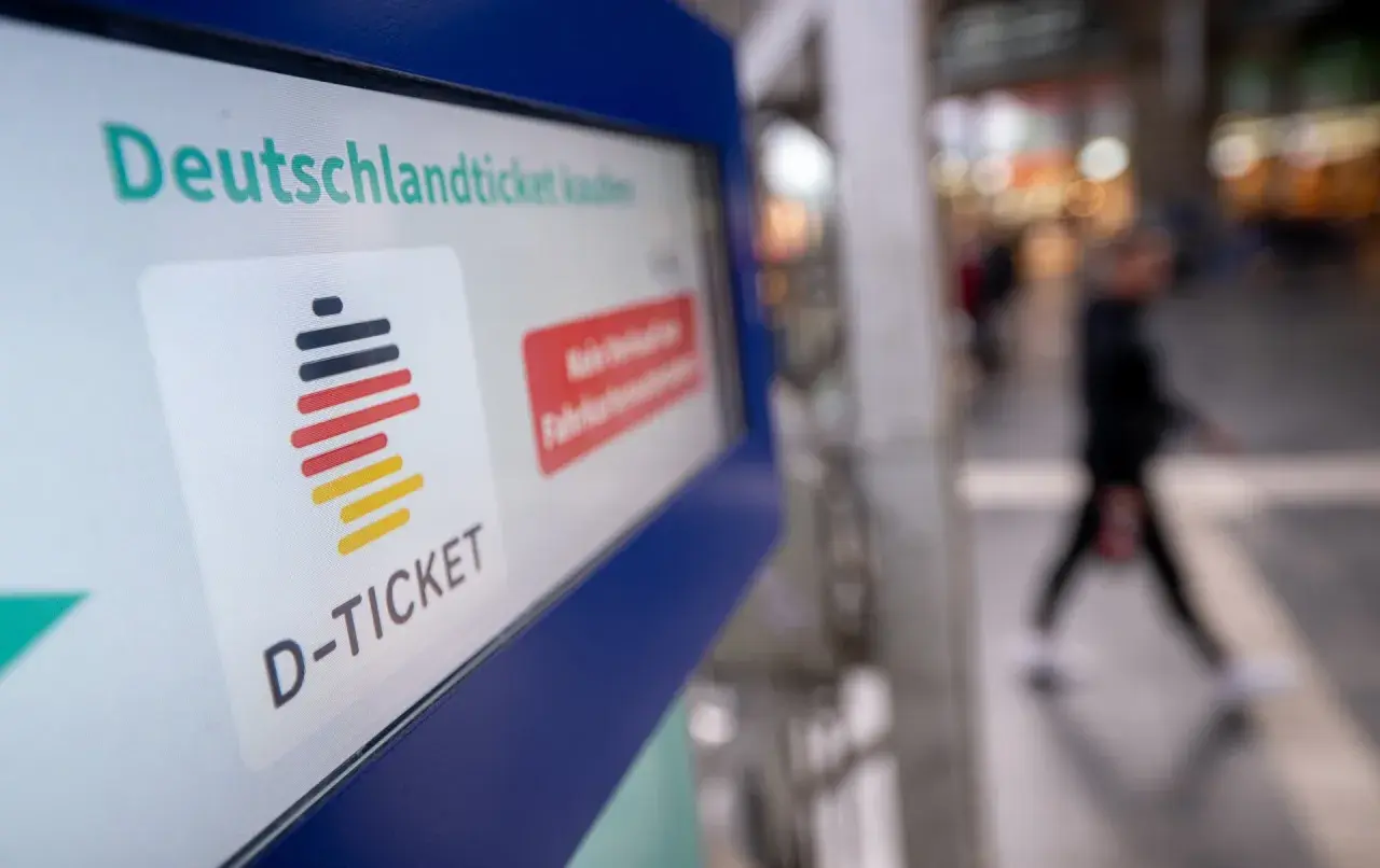 Das Deutschlandticket-Logo auf einem Bildschirm. Eine Person geht unscharf im Hintergrund vorbei. Vielleicht kann man das Deutschlandticket pausieren.