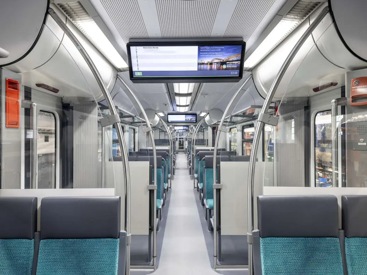 Innenraum eines modernen Zuges mit blauen Sitzen und einem Bildschirm, der die S-Bahn Köln zeigt. Die et 424 ist bereit für die nächste Fahrt.