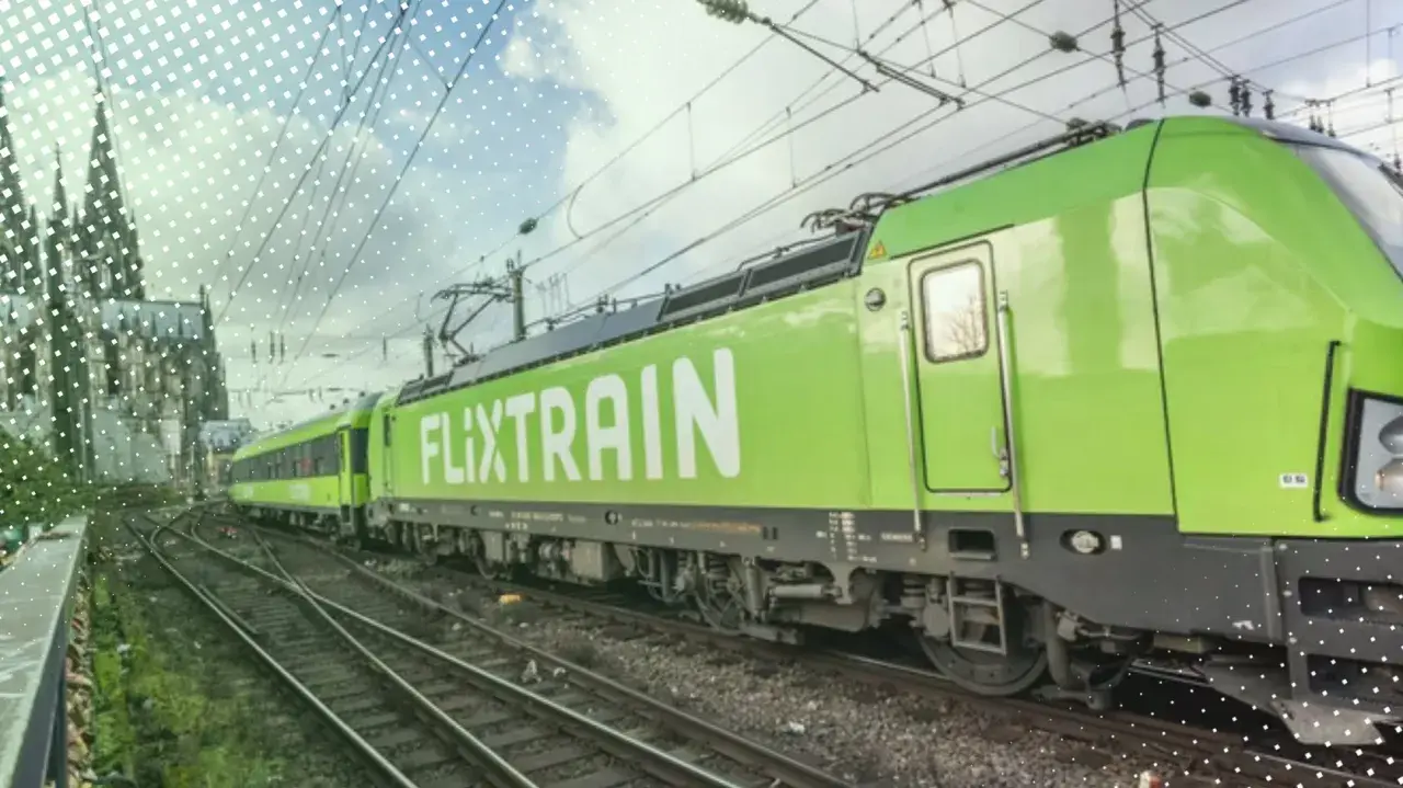 Ein grüner FlixTrain fährt auf Gleisen, im Hintergrund ist eine Kathedrale zu sehen. Perfekt für eine Reise mit dem Deutschlandticket.