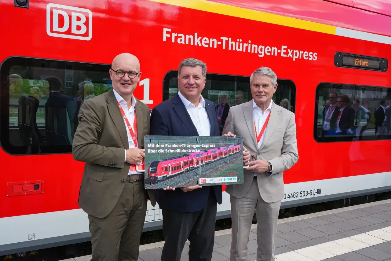 Drei Männer stehen vor einem roten Zug mit der Aufschrift "Franken-Thüringen-Express". Sie halten ein Schild mit einem Bild des Zuges.