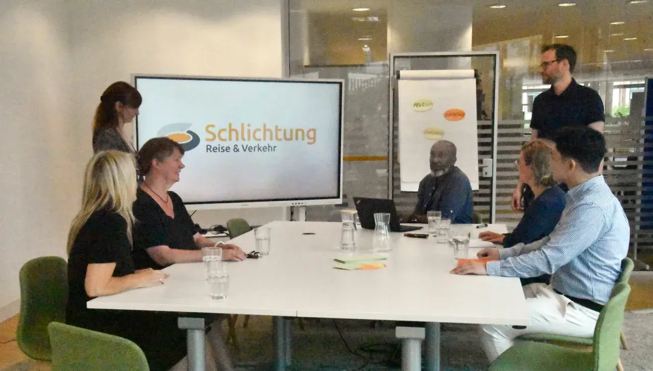 Besprechung im Büro der Schlichtungsstelle Bahn. Mehrere Personen sitzen an einem Tisch, auf dem Bildschirm ist das Logo "Schlichtung Reise & Verkehr" zu sehen.