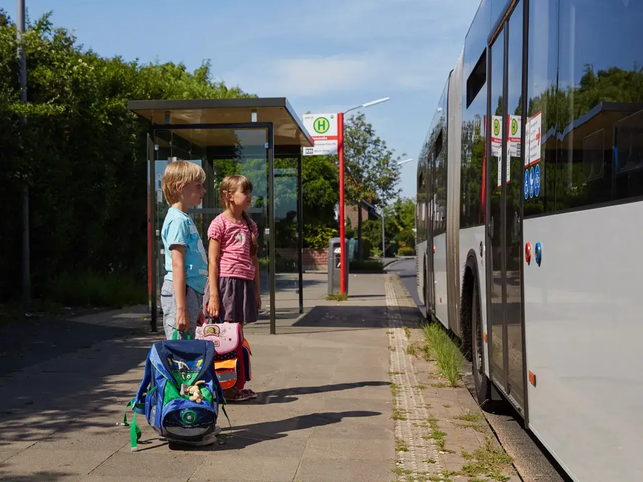 Zwei Kinder warten an der Bushaltestelle auf den Bus. Sie haben bunte Schultaschen dabei.