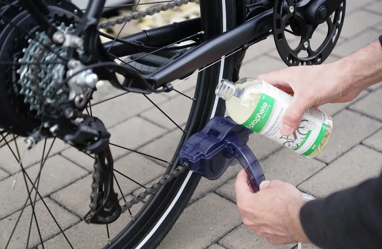 Fahrradkette reinigen mit speziellem Entfetter. Ein Werkzeug reinigt die Kette, während eine Hand die Flüssigkeit aus einer Flasche aufgießt.