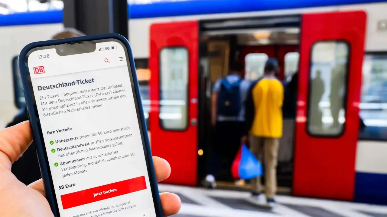 Hand h&auml;lt Smartphone mit App, die das Deutschland-Ticket anzeigt. Im Hintergrund f&auml;hrt eine Bahn ein.