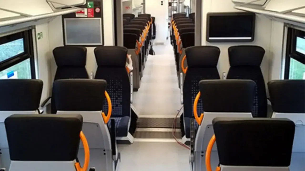 Innenraum eines modernen RRX Zuges mit Reihen von schwarzen Sitzen und orangefarbenen Haltegriffen.