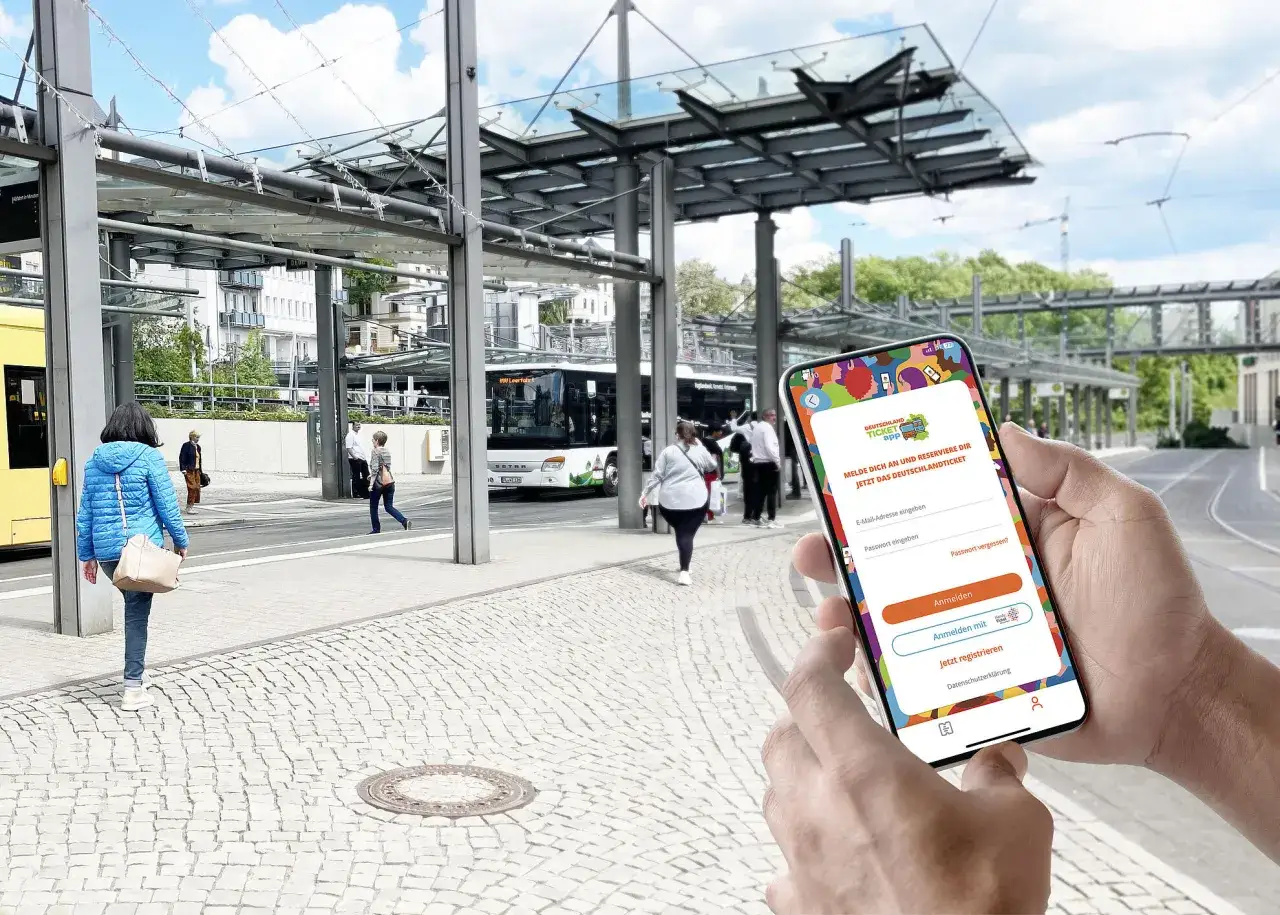 H&auml;nde halten Smartphone mit App zum Deutschlandticket aktualisieren. Im Hintergrund eine Bushaltestelle mit wartenden Menschen.
