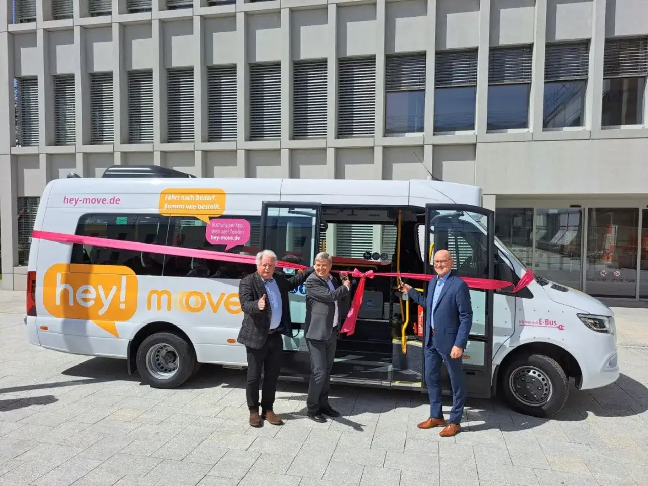 Feierliche Er&ouml;ffnung eines neuen E-Busses f&uuml;r den on demand Verkehr. Drei M&auml;nner durchschneiden ein rotes Band vor dem 