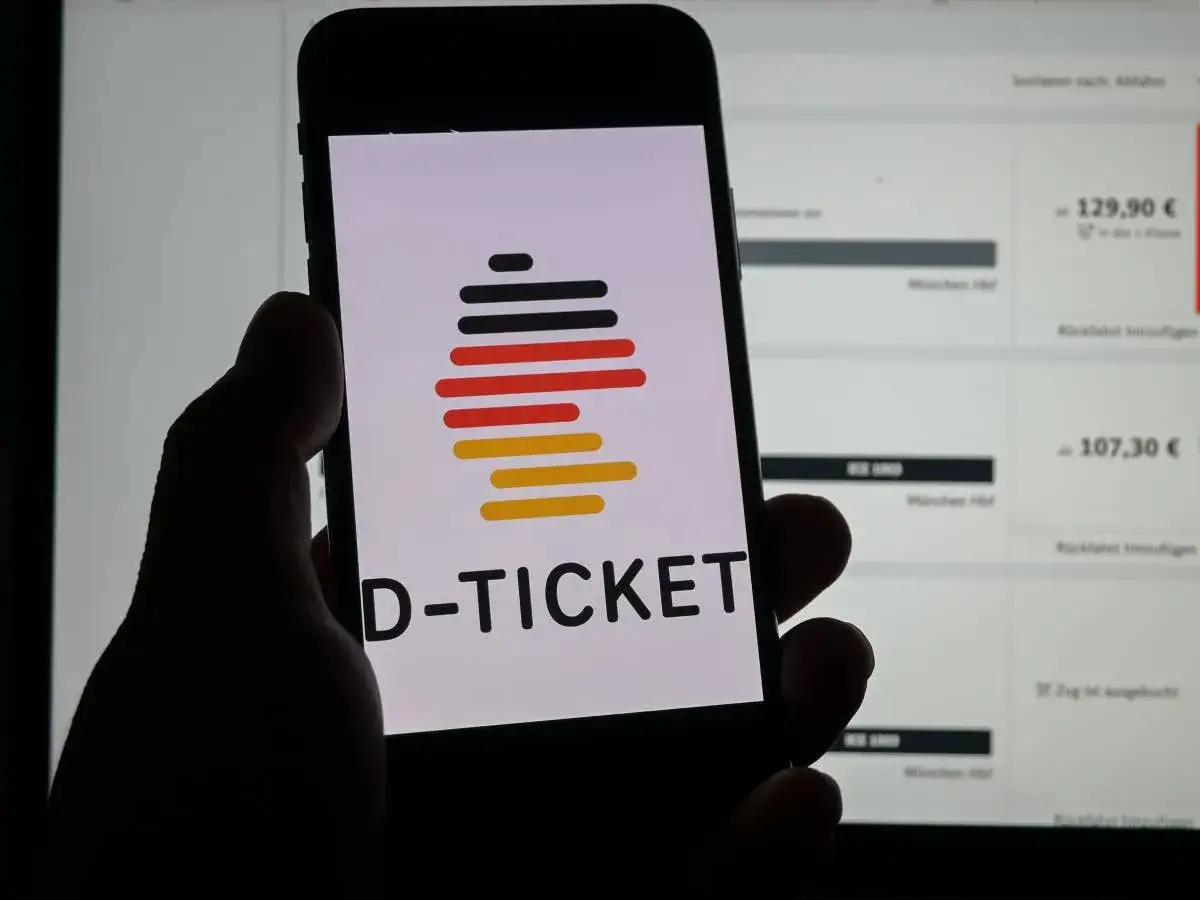 Hand h&auml;lt Smartphone mit D-TICKET Logo. Im Hintergrund Zugtickets, vielleicht das Deutschlandticket nach dem 10. gekauft.
