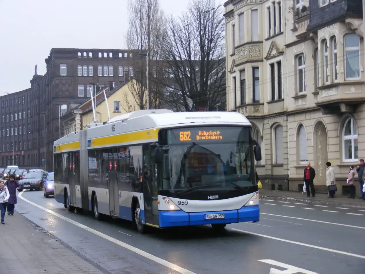 Ein Gelenkbus der Linie 682 f&auml;hrt auf nasser Stra&szlig;e. Menschen warten an der Haltestelle.