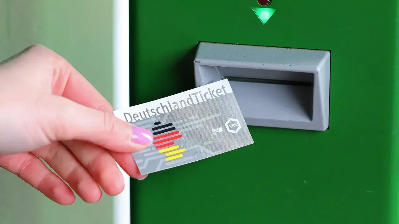 Hand h&auml;lt Deutschlandticket mit VRR-Logo vor gr&uuml;ne Ticketmaschine.