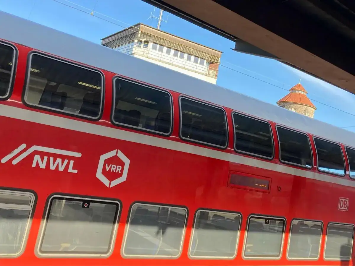 Roter Doppelstockzug mit NWL/VRR-Logo. Im Hintergrund ein Turm, der an eine Ausschreibung von Bahnstrecken erinnert.