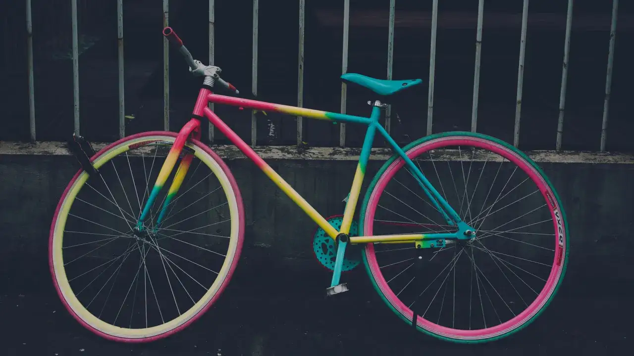 Ein bunt lackiertes Fahrrad mit pinken, gelben und t&uuml;rkisen Akzenten steht vor einem dunklen Gitter.