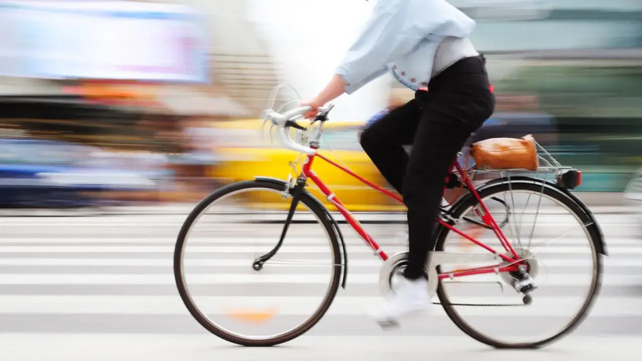 Person auf einem roten Fahrrad f&auml;hrt mit hoher Geschwindigkeit &uuml;ber eine Zebrastreifen. Die Bewegung ist durch den unscharfen Hintergrund und die vorbeiziehenden Autos sp&uuml;rbar.