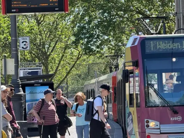 Menschen warten an Gleis 3 auf die Bahn. Die Anzeige zeigt "1 Weiden", was die einfache Fahrt Bedeutung für die Reisenden verdeutlicht.